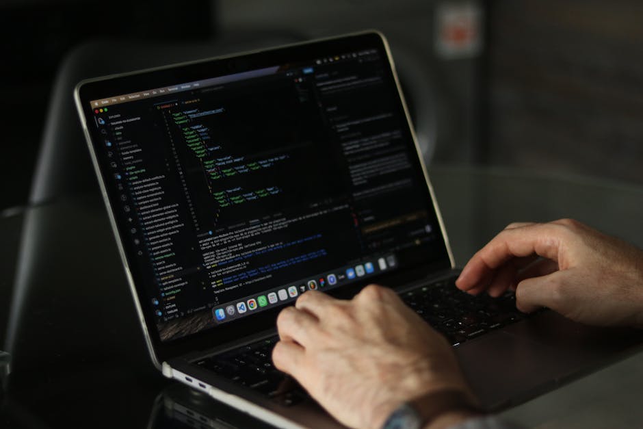 A close-up of a person's hands typing on a laptop with a dark screen displaying lines of code, placed on a black surface. The background is blurred, focusing attention on the device and hands, with so