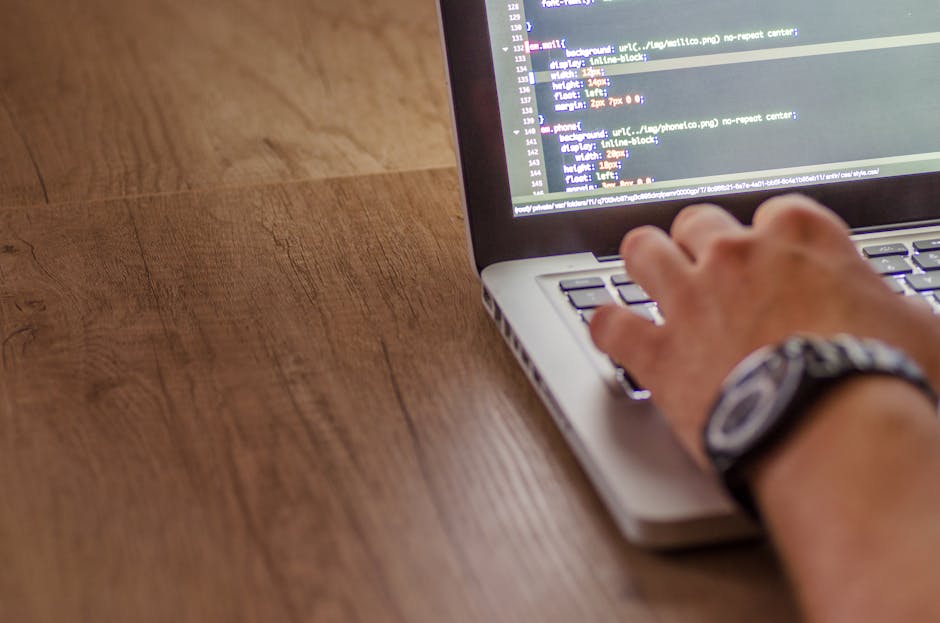 A close-up of a person's hands typing on a laptop with a dark screen displaying lines of code, placed on a black surface. The background is blurred, focusing attention on the device and hands, with so