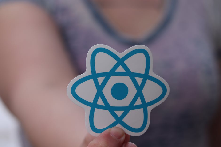 A person holding a sticker featuring a blue and white atom symbol, with a blurred background of a garden scene. In the garden, there are neatly maintained grass lawns with a textured, mowed surface, g